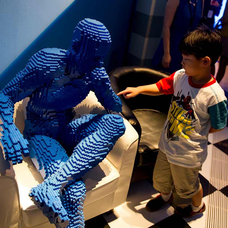 a boy touching a lego sculpture of seated man by Nathan Sawaya